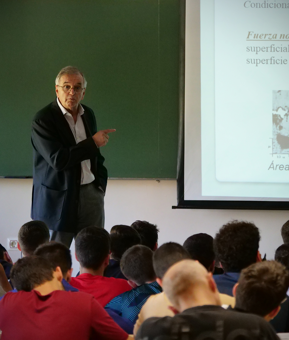 Profesor impartiendo clase con transparencias en aula con estudiantes de espaldas en la Facultad de Ciencias del Deporte