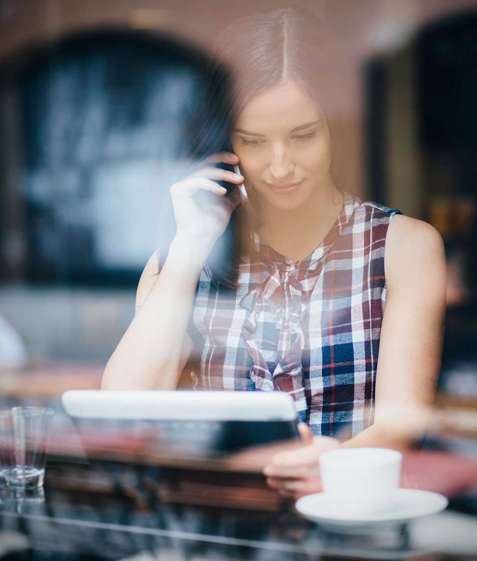 Mujer en el interior de una cafetería tomando café mientras habla por teléfono y lee la pantalla de su tablet