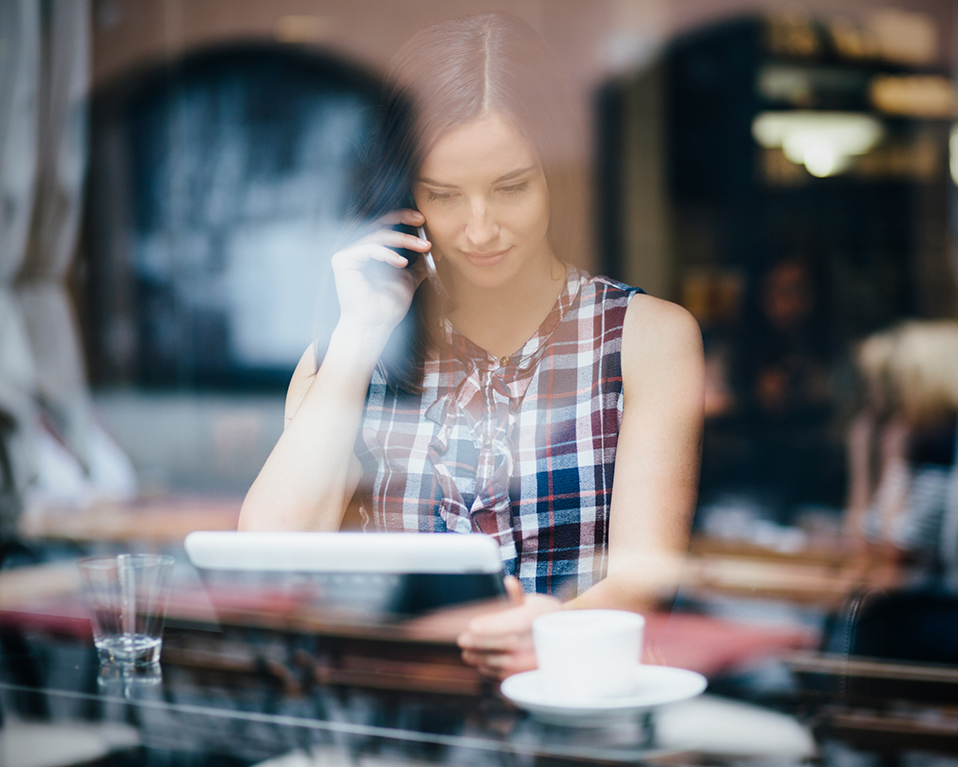 Mujer en el interior de una cafetería tomando café mientras habla por teléfono y lee la pantalla de su tablet